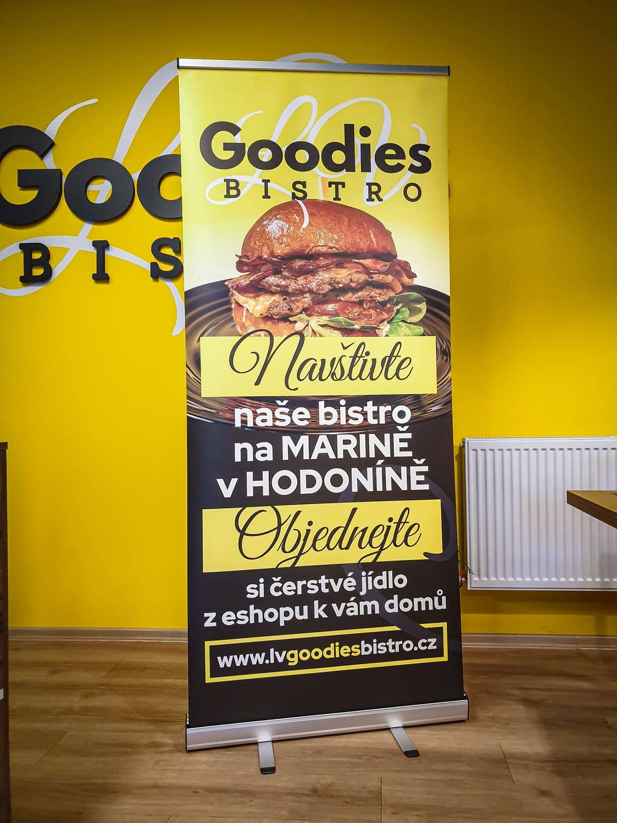 Roll-up Goodies Bistro s burgerom a pozvánkou do bistra pred žltou stenou s 3D logom