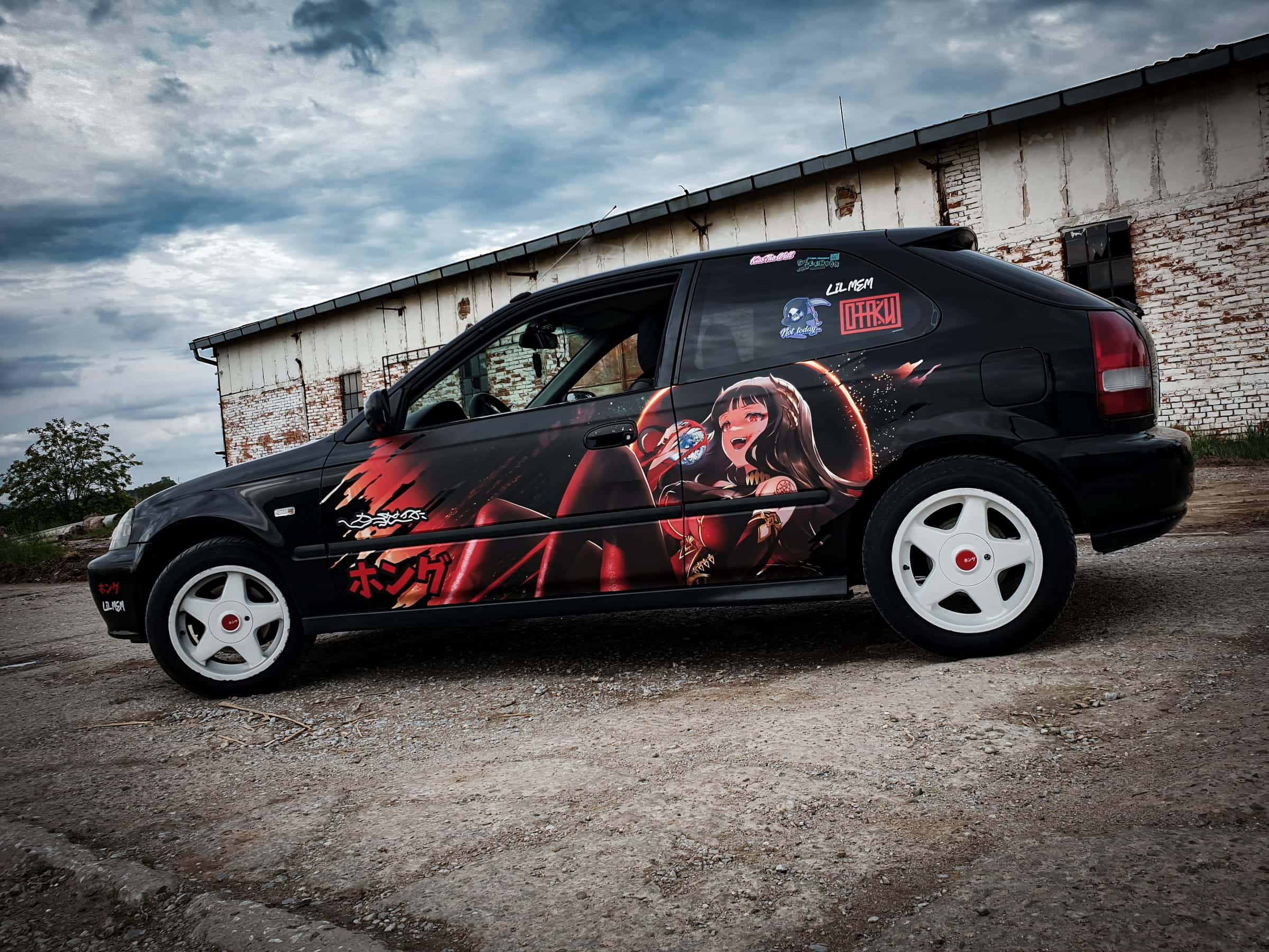Čierna Honda Civic s anime polepom dievčaťa na boku pod dramatickou oblohou