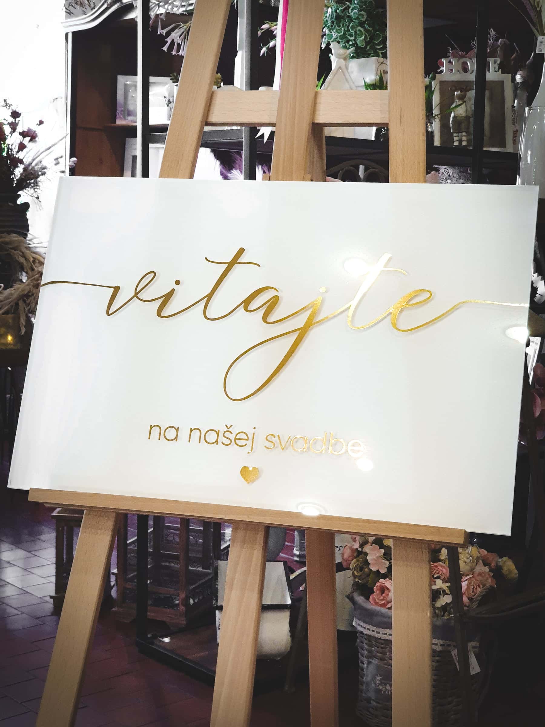 Biela plexi tabuľa so zlatým nápisom Vitajte na našej svadbe na drevenom stojane