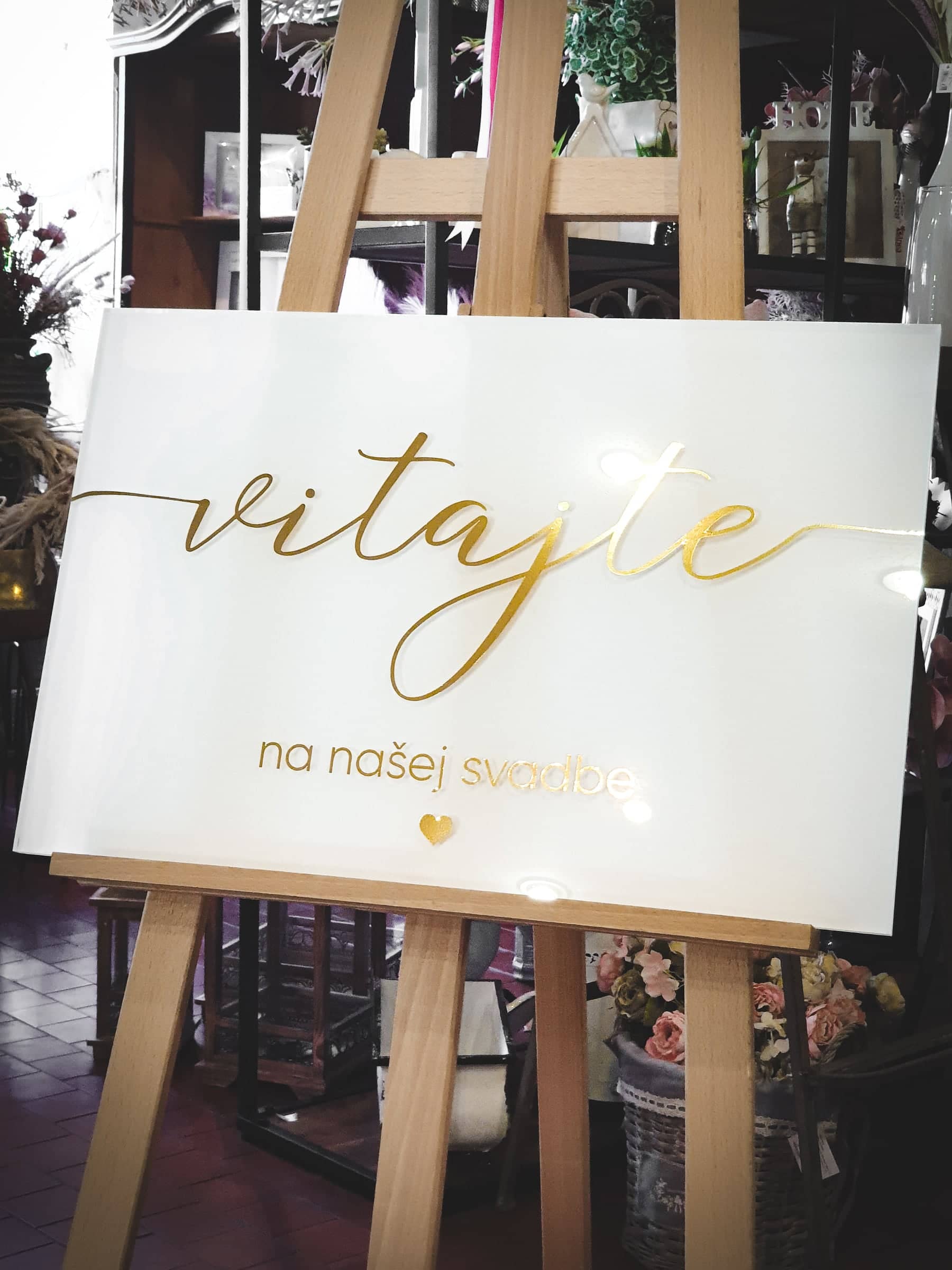 Biela plexi tabuľa so zlatým nápisom Vitajte na našej svadbe na drevenom stojane