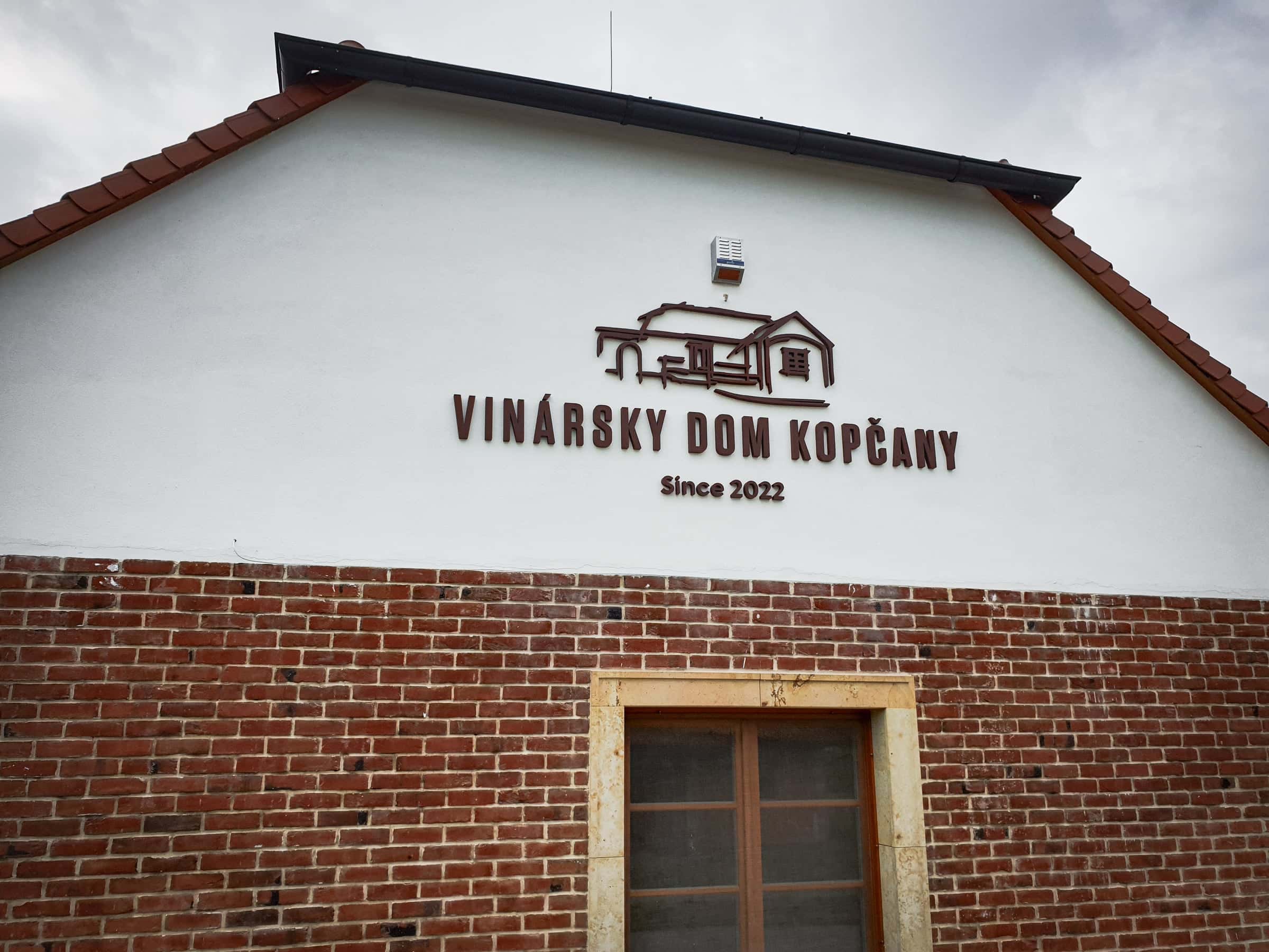 Priestorové 3D logo Vinársky dom Kopčany na bielom štíte budovy s tehlovou fasádou