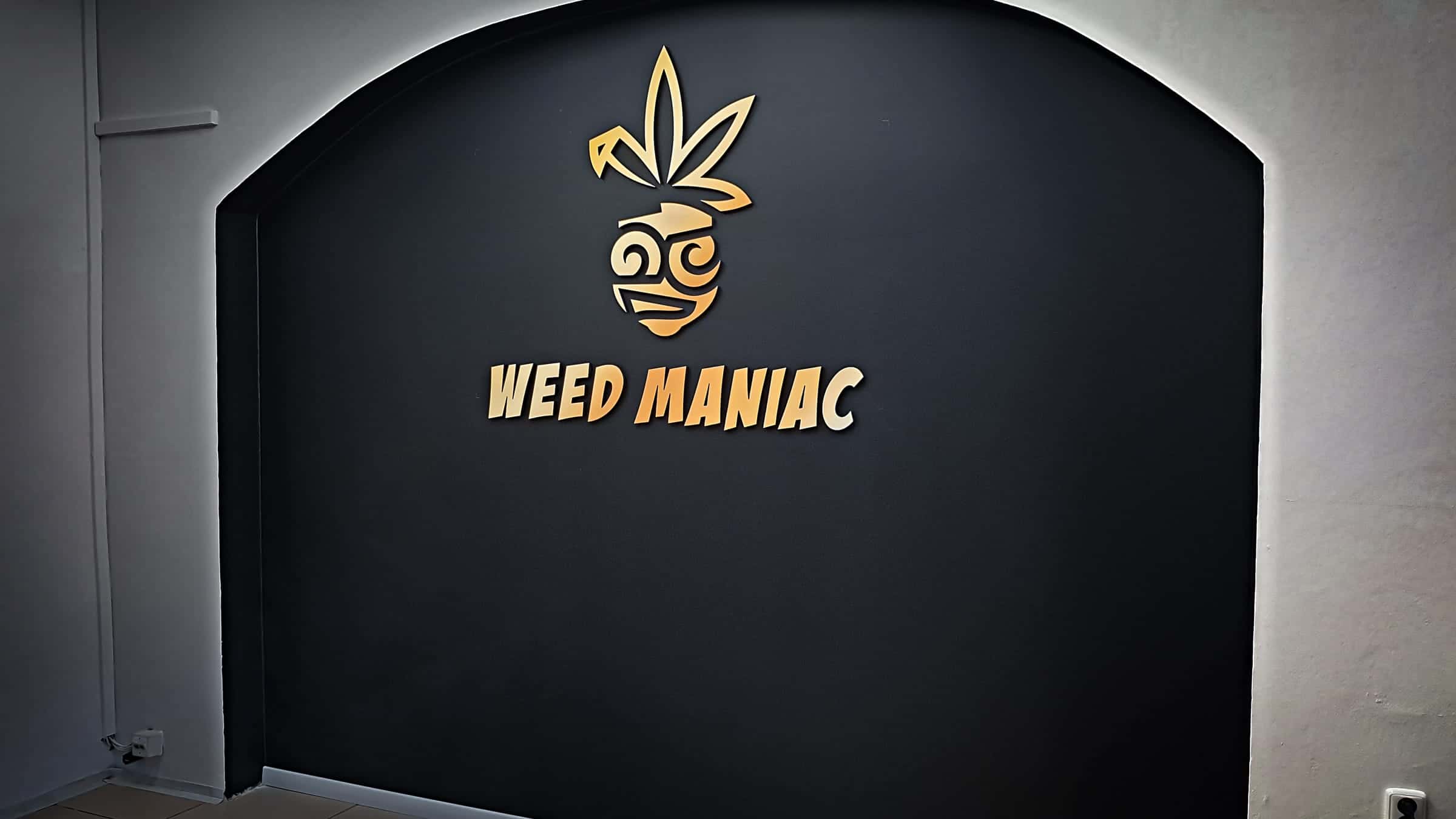 Zlaté 3D logo Weed Maniac na čiernej oblúkovej stene s podsvietením